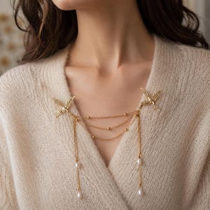Può includere: Una collana color oro con chiusure a forma di uccello e catene pendenti. La collana presenta tre catene con piccole perline dorate e due catene più lunghe con finte perle pendenti. La collana è indossata su un maglione color crema.