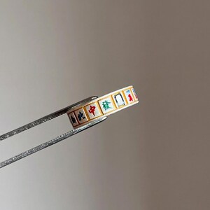 May include: A gold-coloured ring featuring a repeating pattern of colourful Chinese characters. The ring is held by silver tweezers against a neutral background. The characters are in red, green, blue, and black.