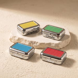 May include: Four rectangular metal tins with hinged lids, each featuring a colored top with a perforated dot pattern. The tins are personalized with the names Seraphina, Dimitri, Aurora, and Leon engraved in a script font on the front side.