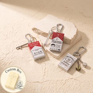 Peut inclure: Trois porte-clés rectangulaires en métal avec mousquetons. L'un présente un motif de paquet de cigarettes rouge et blanc avec le texte "Fucked Up Thursday" et "Less Is Smoke", un autre "MIAOMIAO" avec une clé, et le dernier une étiquette Jack Daniel's.