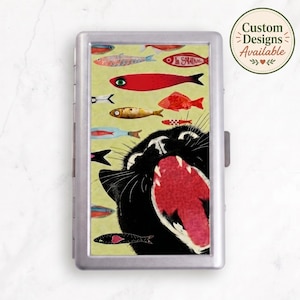Puede incluir: Un estuche rectangular de metal plateado con una ilustración gráfica de un gato negro con la boca muy abierta, rodeado de varios peces coloridos sobre un fondo amarillo claro. El texto Custom Designs Available está impreso en un círculo.