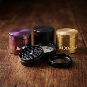 May include: Three cylindrical metal herb grinders in iridescent, matte black, and gold finishes. The iridescent grinder features the text AURORA, while the gold grinder displays GOLDEN ERA. One black grinder is disassembled to show internal teeth.