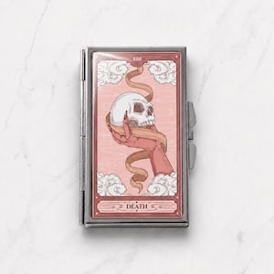 Puede incluir: Un estuche metálico rectangular con una ilustración de carta de tarot en la parte frontal. El diseño muestra una mano sosteniendo un cráneo humano envuelto en una cinta sobre un fondo rosa pálido con nubes. El número romano XIII y la palabra DEATH son visibles.