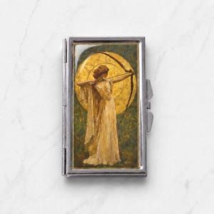 Peut inclure: Un étui rectangulaire en métal argenté présentant une impression décorative incrustée d'une silhouette en robe blanche fluide tenant un arc et une flèche. La silhouette est placée devant un grand fond doré circulaire rappelant le soleil.