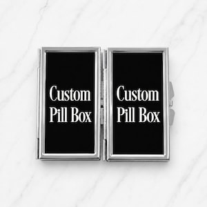 Pode incluir: Uma caixa de comprimidos retangular de metal prateado com design articulado. Os dois painéis frontais apresentam um fundo preto com o texto branco Custom Pill Box impresso em fonte serifada em cada lado. O objeto tem um acabamento metálico polido.