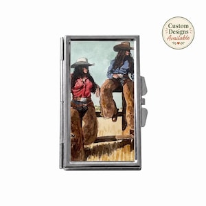 May include: A rectangular silver metal case featuring a watercolor illustration of two figures in cowboy hats, chaps, and western attire sitting on a wooden fence. The top right corner includes a circular badge with the text Custom Designs Available.