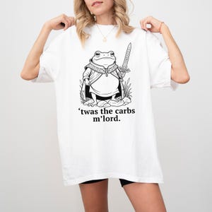 Puede incluir: Camiseta blanca con una ilustración en blanco y negro de una rana vestida como un señor medieval, sosteniendo una espada. El texto "'twas the carbs m'lord." está impreso debajo. Estilo de camiseta casual y unisex.