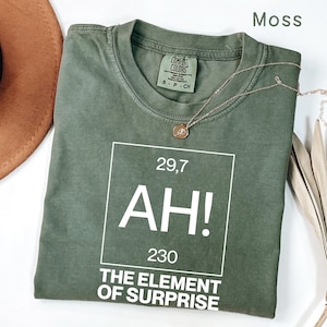 May include: A moss green t-shirt with the text "AH! THE ELEMENT OF SURPRISE" in a white box. A gold necklace with a pendant is draped over the shirt. The shirt is a Comfort Colors brand.