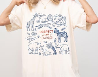 May include: An off-white t-shirt featuring a blue line-art graphic of various safari animals including a giraffe, lion, monkey, zebra, rhino, and elephant. The text "RESPECT THE locals" is printed in the center in dark red and brown lettering.