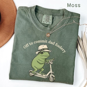 May include: A moss green t-shirt with the phrase "Off to commit dad foolery" and a graphic of a frog wearing a hat and riding a scooter. A silver necklace is draped across the shirt.