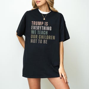 Puede incluir: Camiseta negra con el texto "TRUMP IS EVERYTHING WE TEACH OUR CHILDREN NOT TO BE" en una fuente multicolor. La camiseta la lleva una persona.