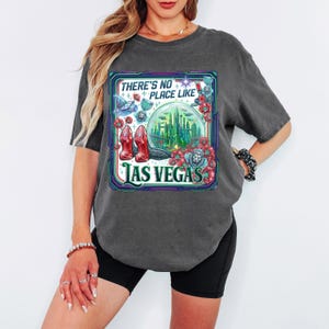 May include: Dark gray t-shirt with a colorful graphic featuring the text "THERE'S NO PLACE LIKE LAS VEGAS." The graphic includes a cityscape, high heels, and floral elements. The shirt has short sleeves.