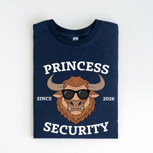 Może przedstawiać: Granatowa koszulka z grafiką byka w okularach przeciwsłonecznych. Tekst "PRINCESS SECURITY" znajduje się nad i pod bykiem, z napisem "SINCE 2026" po obu stronach.