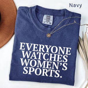 Pode incluir: Camiseta azul marinho com o texto "EVERYONE WATCHES WOMEN'S SPORTS" em letras brancas. Um colar de ouro com um pingente está drapeado sobre a camiseta. A camiseta está dobrada sobre uma superfície branca.