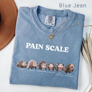 May include: Blue jean colored t-shirt with the text "PAIN SCALE" in white. Below the text is a graphic of opossums illustrating pain levels from "No Pain" to "Worst Pain Possible". A gold necklace is draped across the shirt.