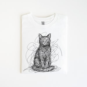 Puede incluir: Camiseta blanca con un diseño de arte de garabatos negro de un gato sentado. El gato está representado con líneas continuas y arremolinadas. El diseño está centrado en la camiseta doblada, que tiene un cuello redondo simple.