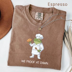 Könnte beinhalten: Ein espressofarbenes T-Shirt mit einer Grafik eines grünen Frosches, der eine weiße Kochmütze und Schürze trägt und ein Brot sowie einen Holzlöffel hält. Der Text "WE PROOF AT DAWN" ist unter dem Frosch gedruckt. Das Shirt hat ein Comfort Colors Etikett.