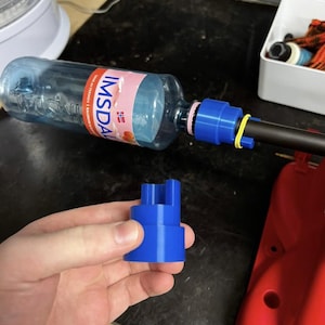 May include: A blue plastic adapter is held in a hand, with a water bottle and a black tube connected to it. The water bottle has a pink and red label. A yellow rubber band secures the tube to the adapter.