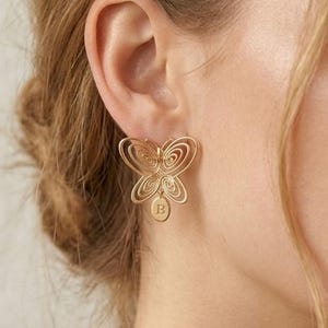 Op de afbeelding: Een goudkleurige vlinder oorbellen met een delicaat, open ontwerp. De oorbel heeft een kleine, ovale bedel met de letter "B" gegraveerd. De vleugels van de vlinder hebben een spiraalpatroon.