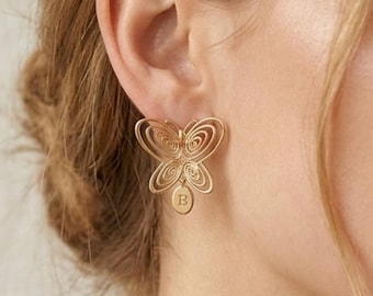 Può includere: Orecchino a farfalla color oro con un design traforato. L'orecchino presenta un piccolo ciondolo ovale con la lettera "B" incisa. Le ali della farfalla hanno un motivo a spirale.