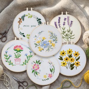 May include: A collection of six white embroidery hoops with floral designs. The hoops feature various embroidered flowers, including lavender, sunflowers, and daisies. One hoop has the text "miss you". The hoops are displayed on a neutral-colored surface.