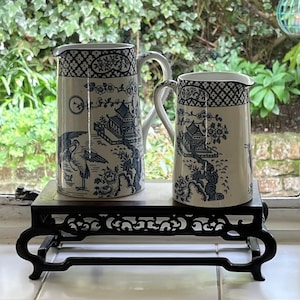 May include: Two ceramic jugs with blue and white Chinoiserie designs, depicting pagodas, birds, and floral patterns. The jugs are on a dark wooden stand with ornate carvings, set against a window.