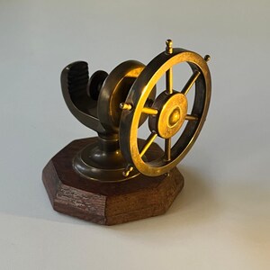 May include: A brass ship's wheel desk accessory with a dark wooden base. The wheel has six spokes and is mounted on a curved support. The base is an octagon shape.