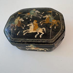 May include: A black octagonal box featuring hand-painted illustrations of figures riding horses and playing polo. The lid and sides are decorated with detailed scenes of riders in colourful clothing, accented by thin white line borders and patterns.