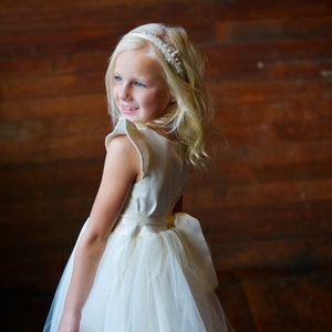 May include: A young girl wearing a white dress with a tulle skirt and a pearl headband. The dress has a satin bodice and a bow at the waist. The girl is standing in front of a wooden wall.