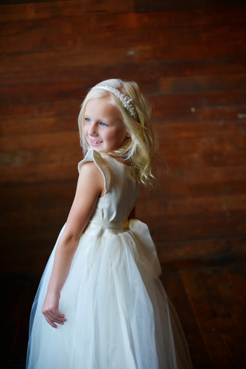 May include: A young girl wearing a white dress with a tulle skirt and a pearl headband. The dress has a satin bodice and a bow at the waist. The girl is standing in front of a wooden wall.