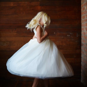 May include: A young girl wearing a white tulle dress with a ruffled neckline, twirling in front of a wooden wall.