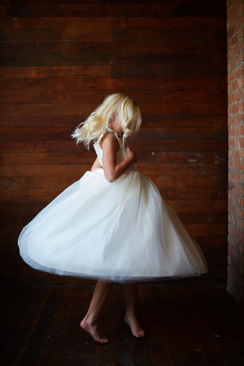 May include: A young girl wearing a white tulle dress with a ruffled neckline, twirling in front of a wooden wall.
