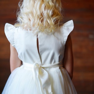 May include: A white tulle dress with a white satin bodice and a bow at the back. The dress has ruffled sleeves and a back zipper.