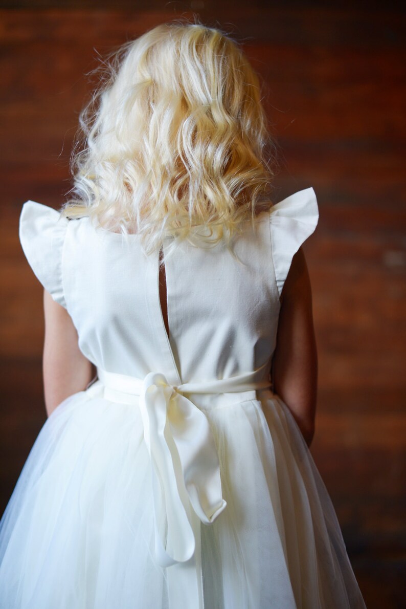 May include: A white tulle dress with a white satin bodice and a bow at the back. The dress has ruffled sleeves and a back zipper.