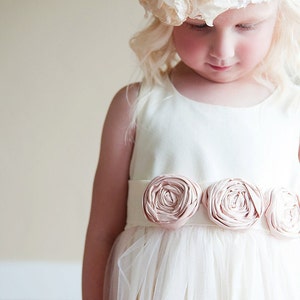 An ivory flower girl dress with a tulle over skirt and blush pink roses on the sash.