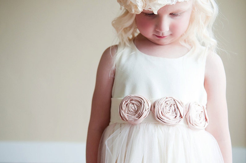 An ivory flower girl dress with a tulle over skirt and blush pink roses on the sash.