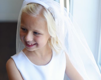 May include: A young girl wearing a white dress and a white veil with a rhinestone headband. She is smiling and looking to the side.