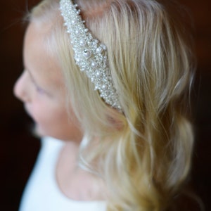 May include: A young girl with blonde hair wearing a white dress and a pearl and rhinestone headband.