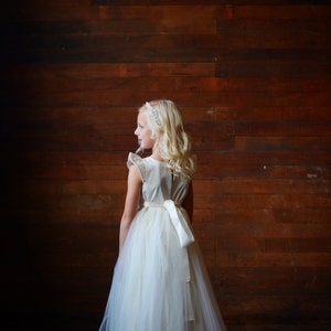May include: A young girl wearing a white tulle dress with a satin sash and a beaded headband. She is standing in front of a wooden wall.