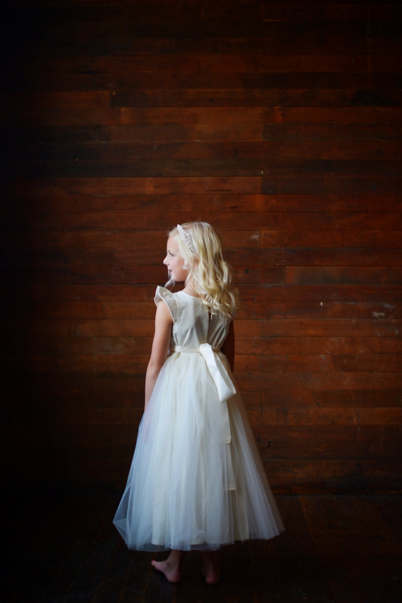 May include: A young girl wearing a white tulle dress with a satin sash and a beaded headband. She is standing in front of a wooden wall.