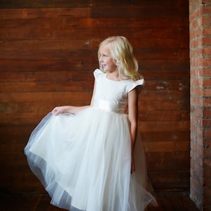 May include: A young girl wearing a white tulle dress with a satin bodice and a white satin sash. She is standing in front of a wooden wall with a brick wall to the right.