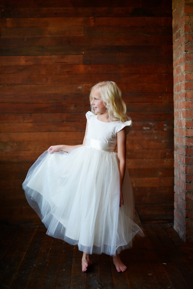 May include: A young girl wearing a white tulle dress with a satin bodice and a white satin sash. She is standing in front of a wooden wall with a brick wall to the right.