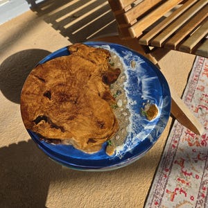 May include: A round wooden table with a blue resin ocean design. The table features a natural wood grain pattern on one side and a detailed ocean scene with sand, rocks, and foamy waves on the other. The table is approximately 18 inches in diameter.