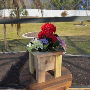 May include: A small, wooden planter box filled with colorful artificial flowers, including red, pink, purple, and white blooms. The planter sits on a round, wooden table with a folding base, set outdoors with a green lawn and trees in the background.