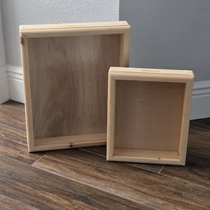 May include: Two rectangular, unfinished light-colored wooden shadow boxes with a natural grain finish. The boxes feature a deep frame design with a slot at the top edge, showcasing a simple, clean construction made from light-toned wood.