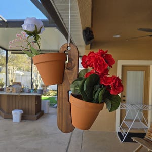 May include: A hanging wooden planter with two terracotta pots, each holding colorful flowers. One pot has white and pink flowers, the other has vibrant red blooms. The planter is suspended by a white string.