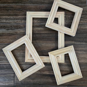 May include: A collection of four unfinished light-colored wooden picture frames with a beveled edge design. The frames are arranged in an overlapping pattern, featuring a natural wood grain texture and a smooth, matte, unpainted surface finish.