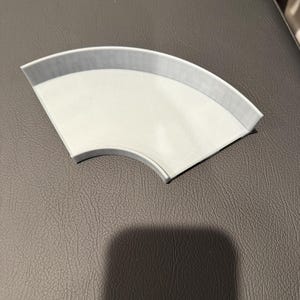 Op de afbeelding: Een lichtgrijs, waaiervormig object met een gebogen rand en een plat oppervlak. Het object lijkt gemaakt te zijn van een vast materiaal en rust op een donkergrijs oppervlak. De vorm suggereert dat het een onderdeel kan zijn van een grotere structuur of ontwerp.