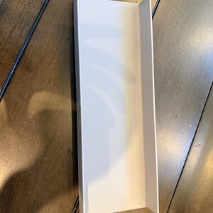 May include: A white, rectangular, U-shaped channel or tray with a flat base and two perpendicular side walls. The object features a smooth, matte finish and a consistent, uniform structure throughout its entire length, showing clean, sharp edges.