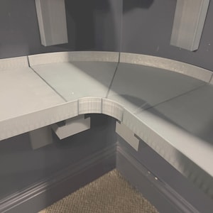 Puede incluir: Una estantería gris en forma de esquina con un borde curvo y un diseño en ángulo recto. La unidad está montada en una pared gris oscuro, con una pieza de soporte a juego visible. La estantería parece estar hecha de un material sólido.